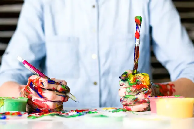 A child with colorful finger paint on both hands sitting at table, engaged in messy play, while holding paint brush and pencil.