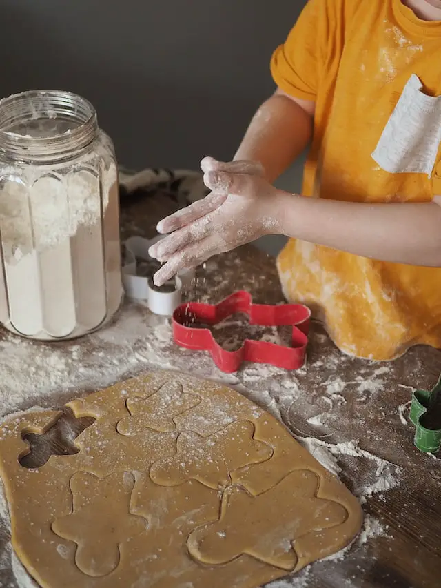 Child making gingerbread cookies. Child is standing at the able with his his hand in flour. Jar of white flour is on table, along with 3 gingerbread cookie cutters (white, red, and blue) and flatten cookie dough. 