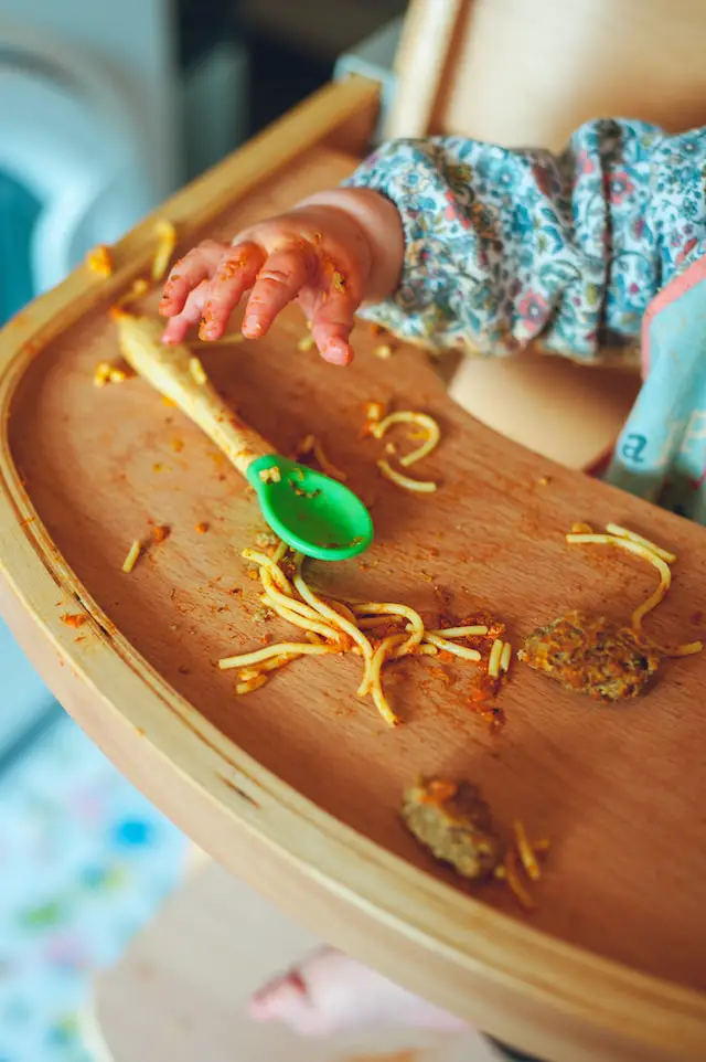Baby's hand messy from spaghetti noodles, sauce and part of meatball, seated in a high chair with tray. Messy food on the tray along a baby spoon.